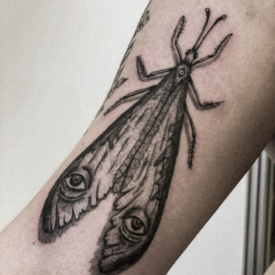 Moth-like tattoo with eyes on its wings and shadows so it seems it is sitting on client´s arm.